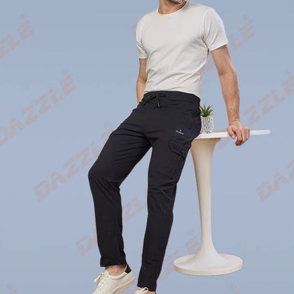 Man wearing sunglasses and black pants standing on a white stool against a light blue background.